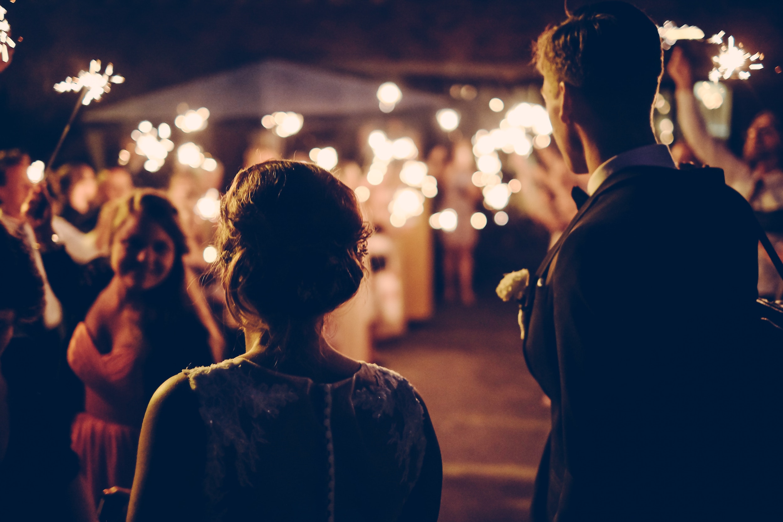 A bride and groom stand facing a joyous crowd of guests holding up lit sparklers at an evening outdoor wedding reception, creating a warm and festive atmosphere.